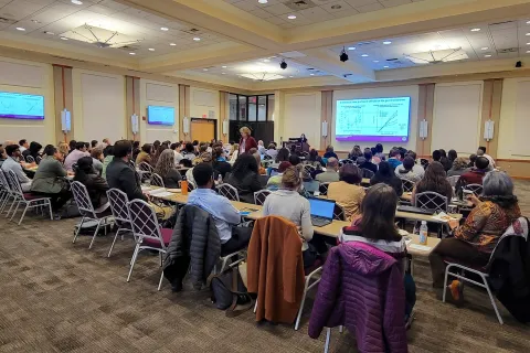 A large audience attends a microbiome research seminar, engaging with a speaker presenting on antibiotics' effects on the gut microbiome.